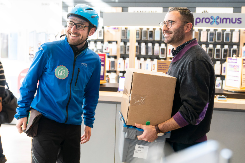 Un trentenaire barbu et à lunettes, employé dans un Proximus Shop, porte deux boîtes - une boîte en carton et une boîte réutilisable en plastique. Il semble qu'il vient de les recevoir d'un coursier en tenue de cycliste sportif avec veste bleu foncé, casque bleu clair et lunettes . Les deux jeunes hommes affichent un large sourire et regardent une troisième personne à l'extérieur de l'image.