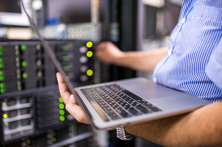 Closeup of a laptop in the hand of a technician. With his other hand, he is reaching for technical equipment that can be seen blurry in the background.