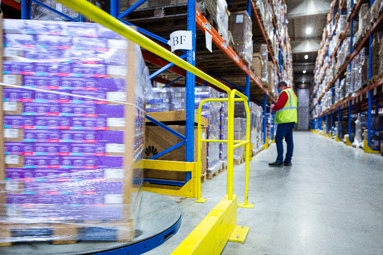 Een magazijnmedewerker zoekt een bestelling tussen metalen rekken. Op de voorgrond staat een pallet met goederen ingepakt op een palletwikkelaar.