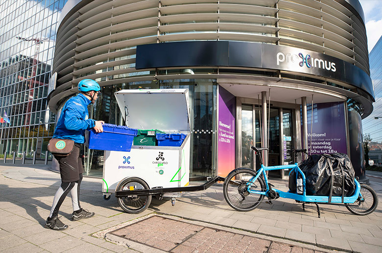 A delivery guy in biking gear and light blue helmet unloads merchandise from the trailer attached to his bicycle. He's dropping it off at a Proximus Store.
