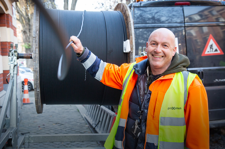 Foto van een straatwerf. Een kale arbeider in fluo vest trekt kabel van een kabelhaspel en kijkt daarbij lachend in de camera.